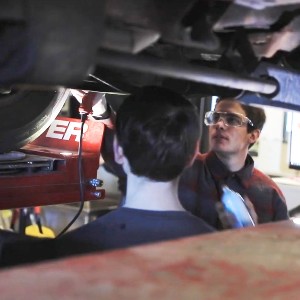 Two students and an instructor inspect the underside of a car.